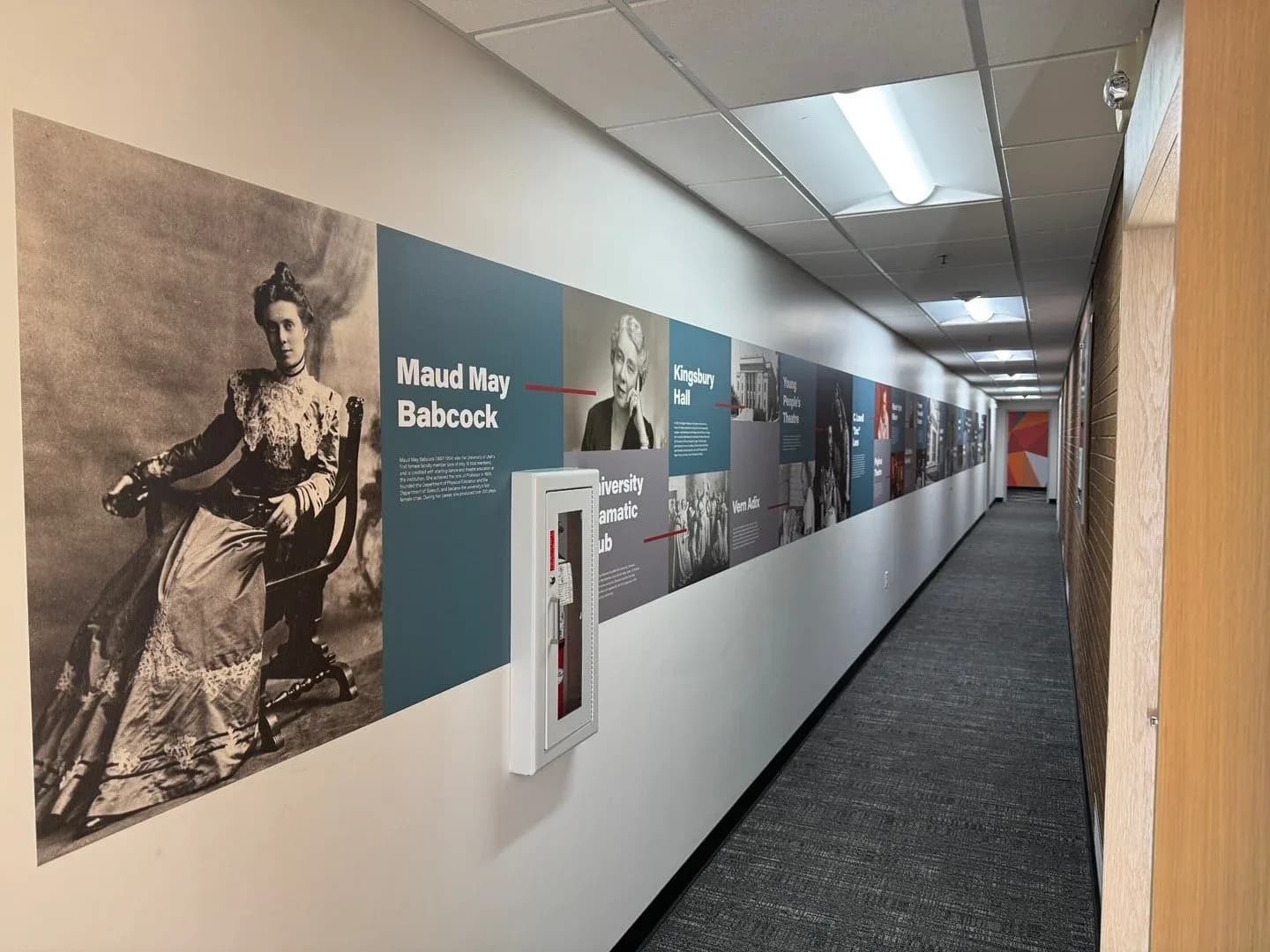 A long hallway with a printed timeline along one wall: a large sepia portrait of Maud May Babcock, then teal and gray panels of photos and text running toward the far end.
